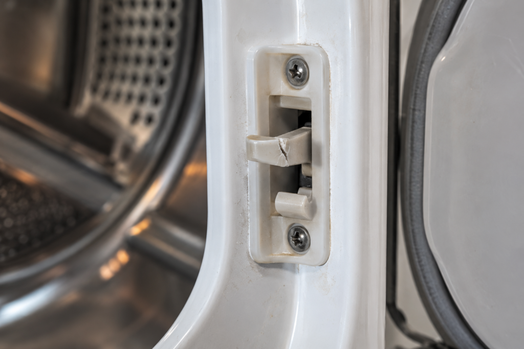Close view of the plastic door latch assembly on a dryer's front frame with one cracked tab.