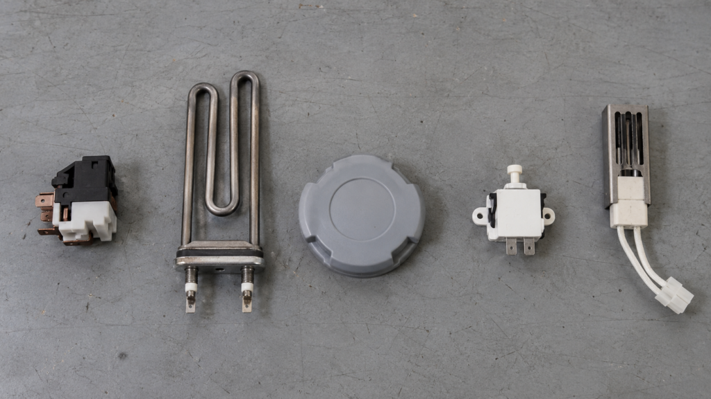 Five small appliance parts laid out in a row on a grey workbench, from a compressor relay to a gas oven igniter.